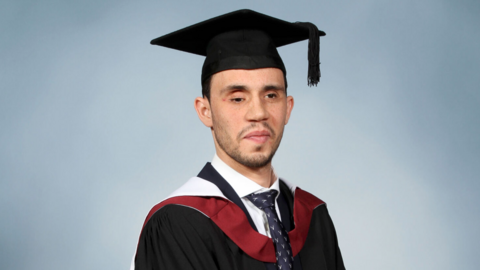 Maher Fattouh in his cap and gown smiling for his graduation photo. He is wearing a black mortarboard, a black robe with red and white accents, a white shirt and a dark blue tie with a white pattern.