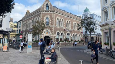 A street corner in a town centre with a large historic brick building featuring arched windows and a central entrance. People are walking, sitting on benches, cycling, and crossing the road, with shops, bicycles, trees, and surrounding older buildings visible under a partly cloudy sky.