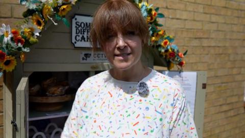 Woman with a colourful top and a fringe stands in front of a cake shed