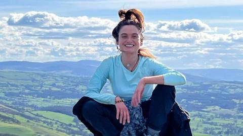Eleanor Thompson is pictured sitting on a rock, with the landscape below, wearing a blue top and jeans, with a jumper tied round her waist. Her hair is tied in a high ponytail and she is smiling.