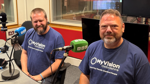 Andreas Findlow and his husband Darren giving an interview in BBC Radio Oxford studio. Darren is looking at the camera and sitting in front of a green microphone. Andreas is sitting in front of a blue microphone.