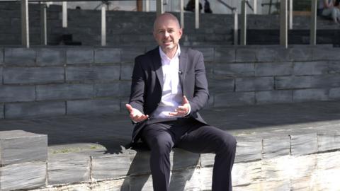 Gareth Lewis sitting on the steps outside the Senedd, wearing a suit and white shirt. He is mid conversation with his hands out in front of him.