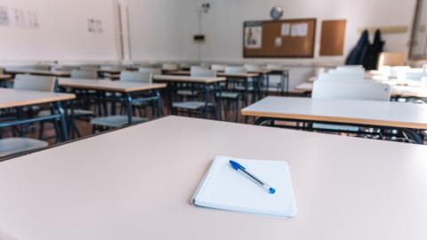 Stock image of a classroom. There are a number of grey desks. The one in the foreground has a notepad and pen on the top.