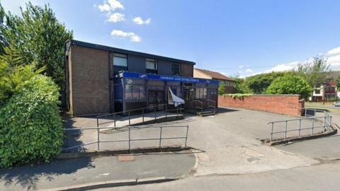 A small two-storey building with a large porch at the front. Some blue signage with indiscernible writing is above the door. A small parking area surrounds the building and it is sunny.