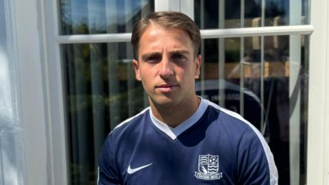 Alexis Cassar looking straight at the camera with a neutral expression. He is standing outside, in front of a window, and is wearing a navy blue football shirt with a white Nike tick on the left and a white Southend United badge.