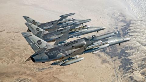 Draken's Alca L-159E "Honey Badger" aircraft flying in formation. Sand covers the ground below them. The three planes are lined up and flying to the right. They are tilted away from the camera as if turning.