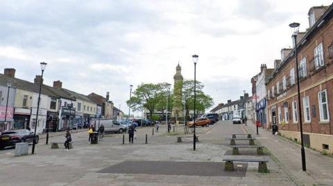Tower Square in Tunstall, with paving and benches as well as a central clock tower. There is car parking in the middle and shops on either side of the public square.