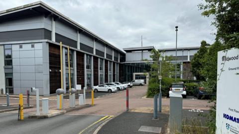 A picture showing a car parking barrier, and a large office block - parked outside are several cars. 