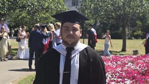 Oliver, who has dark hair and short facial hair, is wearing graduation robes and a cap, standing beside a flower bed in a park, with other graduates in the background.