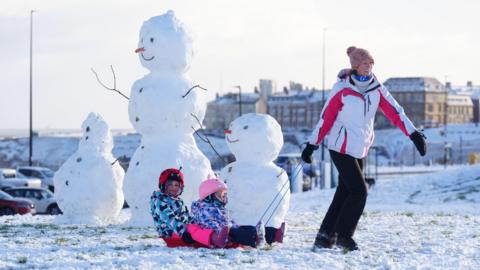 A woman wrapped in warm clothing pulls two children on a sled in the snow. They are both wrapped up in coats and have helmets on. Behind them are three snowmen. The middle snowman is taller than the adult woman in the photo. The two either side of it are around half the height. 