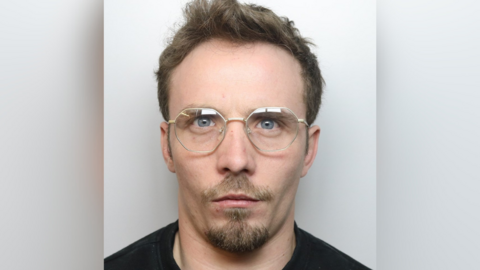 Thomas Harris mugshot. He has a goatee-like beard, moustache and is wearing glasses. He has quiffed brown hair.