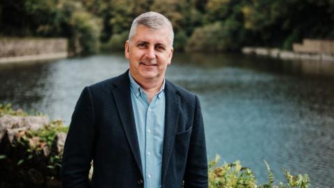 Helier Smith standing in front of a body of water and waist-height stone wall, looking directly into the camera, wearing a smart dark blue jacket with lighter blue button up shirt below