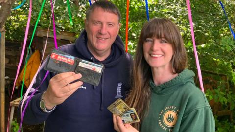 A man in a blue hoodie holding a wallet stood next to a woman in a green hoodie holding some cards which were found in the wallet. Thet are in a woodland with bright coloured streamers in the background. 