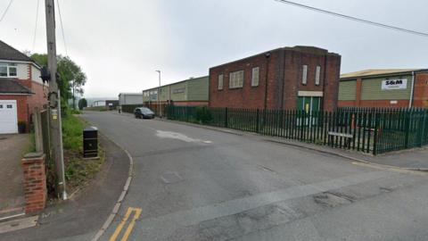 A general view of Canal Lane - a house is on the left and industrial buildings are behind a green fence on the other side of the road with a street sign that says "Canal Lane" in front of the fence.