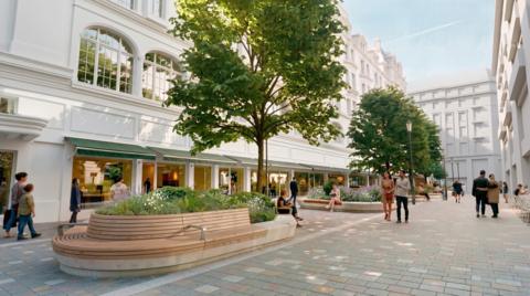 Image of a wide pedestrianised street lined with trees, benches and planted flower beds. People are walking, sitting and socialising outside shopfronts beneath white buildings with green awnings.