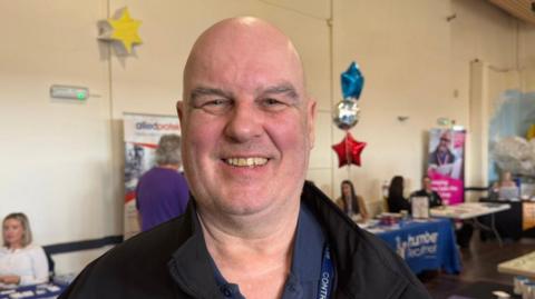 The photo shows a man smiling at the camera. He has a bald head and is wearing a black jacket. He's standing in a room which has a number of tables behind it, advertising businesses. There are three balloons to his right. 