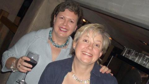 A middle-aged woman with short dark hair and wearing a pale blue cardigan is holding a glass of red wine and smiling at the camera. Her other hand is on the shoulder of another woman of a similar age with shortish blonde hair, who is also smiling at the camera.