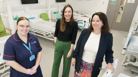 Left to right: SACT Clinical Educator at South Tyneside and Sunderland NHS Foundation Trust (STSFT) Rachel Boyd, STSFT Oncology Nurse Consultant  Lynsey Robson and programme leader Karen Humphreys smiling inside the University of Sunderland’s mock hospital ward. There are two hospital beds behind them with a green chair and a chest of drawers. 
