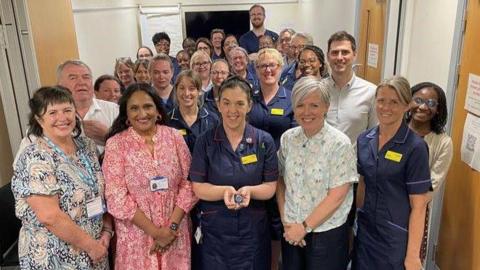 A large group of people smile at the camera with Alison Rea front and centre holding her award, they are standing in a corridor