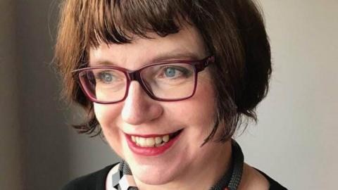 A woman with a brown bob is looking off to the side with a lovely smile. She is wearing glasses. 