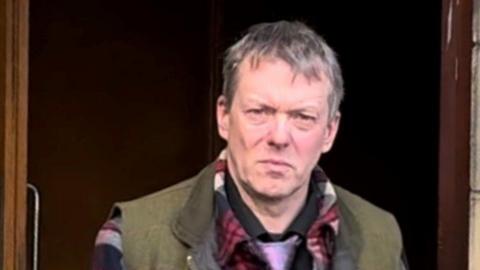 Mark Craster-Chambers is walking out of court, but is looking at the camera. He has short grey hair and a perplexed look on his face. He is wearing a green body-warmer, a red checked shirt and a purple tie.