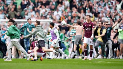 Football fans storm the pitch after Celtic beat Hearts. Two despondent Hearts players can be seen - one crouched on his knees, the other simply walking off. Fans in Celtic shirts are running around, in one case taunting them.
