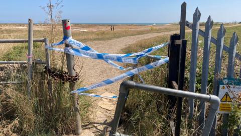 Police tape across a fence blocking off a path to the beach 
