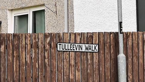 A cropped photo showing a street sign at Tulleevin Walk, Newtownabbey, attached to a brown wooden fence. There is a lamp post to the right of the sign and a building in the background with a white window.