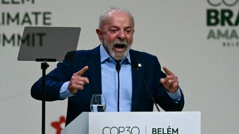 Brazilian President Lula stands behind a white podium with the words "Belém COP30". On the podium is a glass of water. Lula is in a blue suit his hands pointing forward and his mouth open mid-speech.