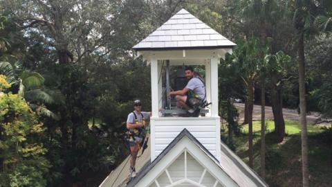 Two men in the chapel's tower installing the bell. They are smiling broadly. Behind the white chapel are trees and a road.