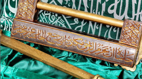 A wooden lock inscribed with Arabic verses from the Quran. It sits on a green cloth.