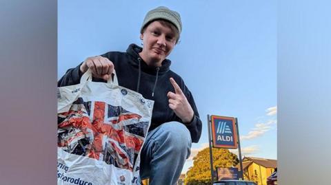 Leroy Lupton kneeling down with Aldi bag of shopping outside an Aldi store 