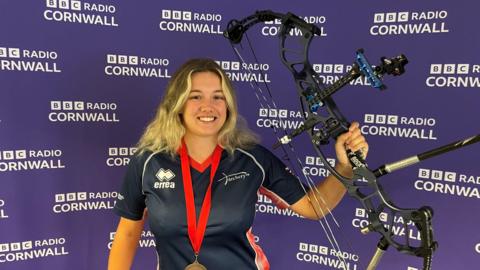 Chloe is smiling at the camera holding her compound bow up high. She is wearing her GB archery uniform and a bronze medal with a red ribbon. She has shoulder-length blonde hair. The purple backdrop is covered with repeated white BBC Radio Cornwall logos. 