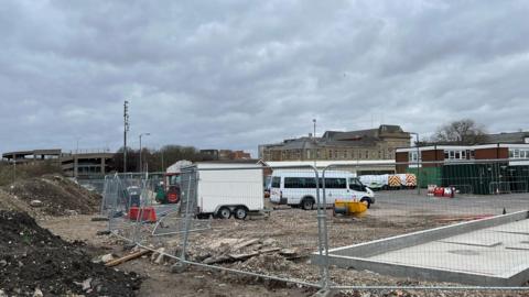 Mounded earth in front of a metal barrier along the perimeter of the site, with vans and a trailer parked behind it