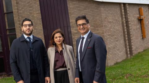 Three representatives of the Malhotra Family Foundation outside the United Reformed Church in Ponteland. Two men, both wearing navy suits and ties, both with dark short hair and glasses, stand at either side of a woman. She has long brown hair and is wearing black trousers, a purple blouse and cream coat. 