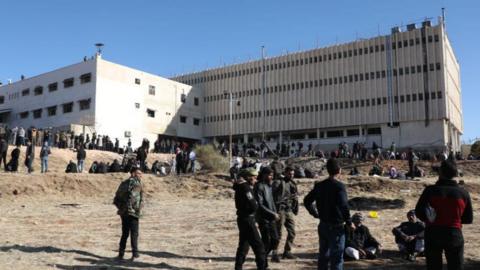 People gathering outside Saydnaya Prison, which is a big white building.