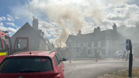 Grey smoke rises from wooden buildings into the sky. Part of a red fire engine is visible.