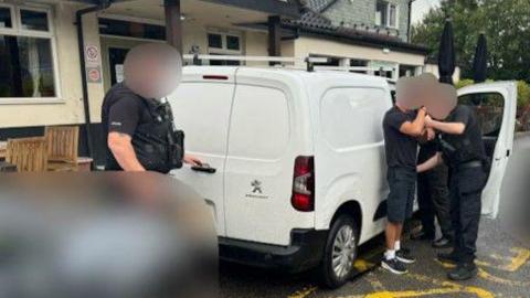 Two police officer can be seen detaining a man by the side of a white van which is parked next to what appears to the the entrance of a pub. Another police officer can be seen with a hand on the handle of the van's read door.