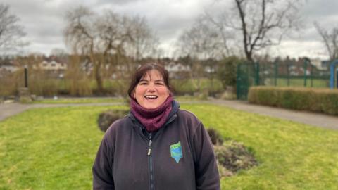 A woman with dark hair is standing in hull road park, there's grass all around her and trees in the distance. To the right of her is the fence to the tennis courts. There's a path just before round the edge of the courts. The woman is smiling, wearing a black fleece and dark purple scarf. 