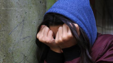 A teenage girl with black hair wearing a blue hooded top and a purple coat puts her hands over her face while in an upset state. She is stood next to a concrete pillar.