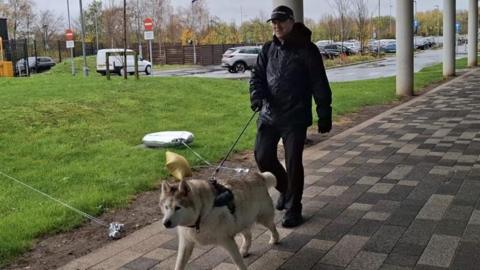 Insp Andy Robinson in a waterproof black coat, police cap and black gloves holding a lead to attached to a white husky dog.