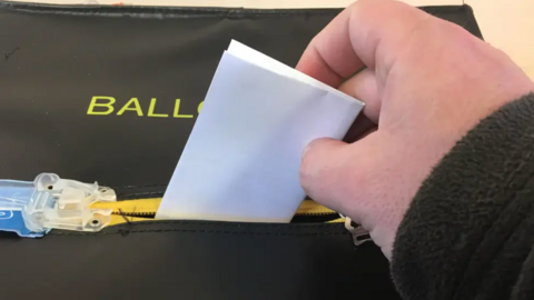 A hand placing a folded piece of paper into a black ballot box.