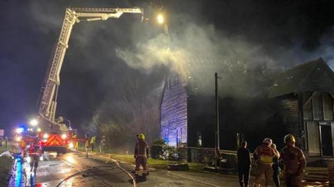 A fire engine and firefighters on a road next to a house.
There is a lot of smoke coming from the house.
The picture was taken in the evening and it is dark outside.