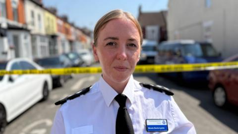 Ch Insp Nicola Davis-Lyons, who is standing in the middle of a street with terraced houses and cars on either side. Her fair hair is pulled back from her face and she is wearing a white police uniform shirt with epaulettes on her shoulders and a black tie. Behind her is a line of yellow police tape.