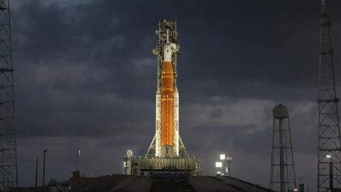 Orange rocket with white tip stands vertically ready to lift off lit up by flood lights