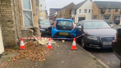 The rear of a blue car is seen wedged between two parked cars. To the left is a house with part of its lower wall destroyed by the car that crashed into it. There are police cones and tape cording off the scene. 