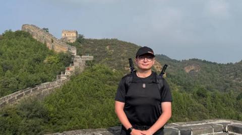 Will Blisset wears a black t-shirt, shorts and cap. He stands in front of a section of the Great Wall of China. You can see lush green trees in the background and a  walk that snakes up into the hills.