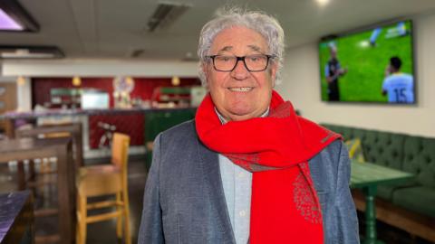 Gilberto smiles at the camera as he wears a grey blue jacket with a light blue shirt underneath and he's also got a long red scarf on as well. He has glasses and grey, curly hair. Behind him are tables and chairs and a screen showing football and at the back of the room there is a bar.