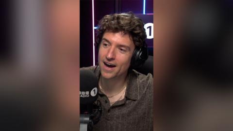 A man in a brown shirt speaks into a radio 1 microphone.