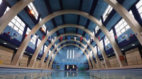 A swimming pool with parabolic arches. the ceiling is painted blue with four dark blue lines down it. There is bunting running across the pool and windows either side